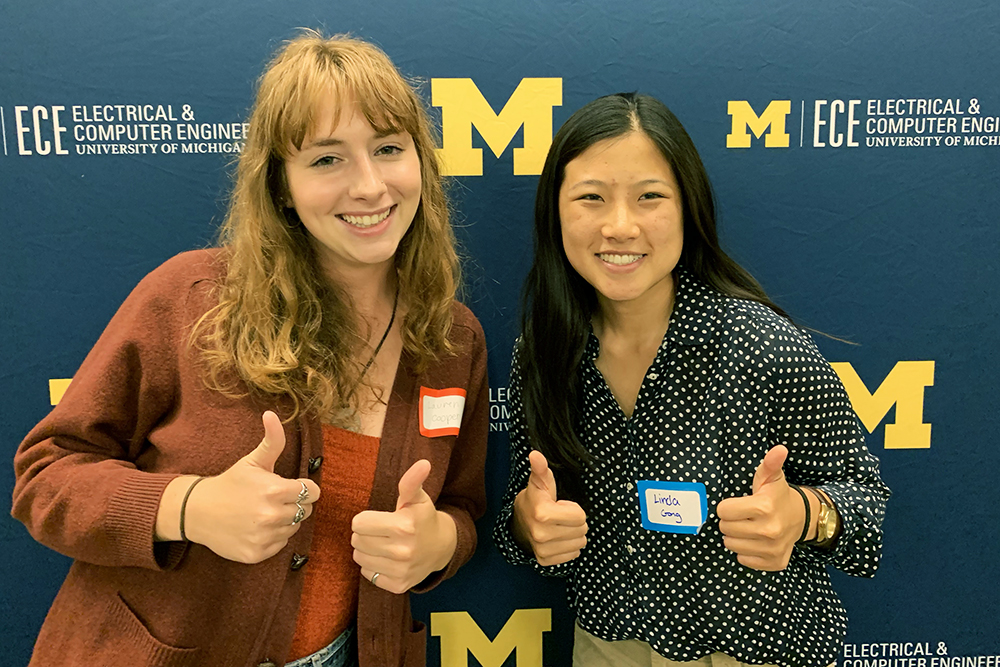 Two women pose with thumbs up in front of U-M ECE backdrop
